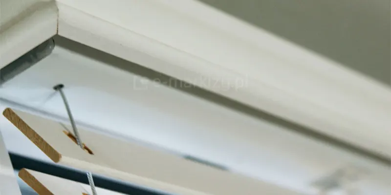 Zdjęcie prezentuje szczegółowy widok na górny mechanizm montażowy oraz fragment lameli, które tworzą żaluzje drewniane 25 mm w kolorze białym. Widoczna jest górna rynna osłonięta estetyczną, białą maskownicą drewnianą o gładkim wykończeniu, która przylega bezpośrednio do ramy okiennej. Poniżej maskownicy znajdują się pierwsze poziome lamele o szerokości 25 mm, ustawione pod kątem, co odsłania ich naturalny, drewniany profil na bocznych krawędziach. Przez precyzyjnie wycięte otwory w listewkach przechodzą cienkie, stalowe linki prowadzenia bocznego, które stabilizują żaluzje drewniane 25 mm przy szybie, zapobiegając ich odchylaniu przy uchylonym oknie. Linki są naprężone i biegną pionowo, przechodząc przez każdą lamelę. Cała konstrukcja utrzymana jest w minimalistycznej, jasnej kolorystyce, gdzie biel lakierowanego drewna dominuje w kadrze, a delikatne akcenty naturalnego odcienia drewna widoczne są jedynie w przekroju listewek. Perspektywa zdjęcia z dołu ku górze podkreśla precyzję spasowania elementów systemu oraz matową teksturę białych komponentów.