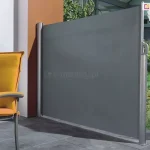 Zdjęcie prezentuje boczną markizę tarasową, pełniącą funkcję parawanu ochronnego, zamontowaną na nowoczesnym tarasie. Osłona składa się z pionowej kasety przymocowanej do ściany budynku oraz wysuwanej tkaniny w kolorze ciemnoszarym o gęstym splocie. Materiał jest całkowicie rozciągnięty i zamocowany do wolnostojącego, srebrnego słupka końcowego przytwierdzonego do podłoża. Na słupku widoczny jest ergonomiczny uchwyt służący do rozwijania i blokowania osłony. Podłoże tarasu wyłożone jest dużymi, kwadratowymi płytami w kolorze antracytowym. Po lewej stronie kadru znajduje się fragment mebla ogrodowego – krzesło z ciemną ramą i podłokietnikami, wyposażone w grube, intensywnie żółte poduszki na siedzisku i oparciu. Tło za parawanem stanowi fragment ogrodu z zieloną trawą, niską roślinnością oraz nowoczesną architekturą budynku z metalowymi elementami konstrukcyjnymi. Po prawej stronie, obok słupka markizy, stoi wysoka, smukła donica w kolorze czarnym z ozdobnym krzewem. Światło słoneczne pada z lewej strony, tworząc wyraźne cienie na płytach tarasowych i podkreślając teksturę materiału osłony.