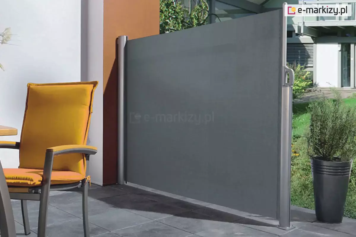 Zdjęcie prezentuje boczną markizę tarasową, pełniącą funkcję parawanu ochronnego, zamontowaną na nowoczesnym tarasie. Osłona składa się z pionowej kasety przymocowanej do ściany budynku oraz wysuwanej tkaniny w kolorze ciemnoszarym o gęstym splocie. Materiał jest całkowicie rozciągnięty i zamocowany do wolnostojącego, srebrnego słupka końcowego przytwierdzonego do podłoża. Na słupku widoczny jest ergonomiczny uchwyt służący do rozwijania i blokowania osłony. Podłoże tarasu wyłożone jest dużymi, kwadratowymi płytami w kolorze antracytowym. Po lewej stronie kadru znajduje się fragment mebla ogrodowego – krzesło z ciemną ramą i podłokietnikami, wyposażone w grube, intensywnie żółte poduszki na siedzisku i oparciu. Tło za parawanem stanowi fragment ogrodu z zieloną trawą, niską roślinnością oraz nowoczesną architekturą budynku z metalowymi elementami konstrukcyjnymi. Po prawej stronie, obok słupka markizy, stoi wysoka, smukła donica w kolorze czarnym z ozdobnym krzewem. Światło słoneczne pada z lewej strony, tworząc wyraźne cienie na płytach tarasowych i podkreślając teksturę materiału osłony.