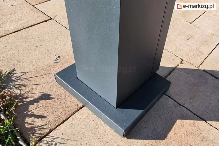 Zdjęcie prezentuje szczegółowe ujęcie dolnej części pionowego słupa konstrukcyjnego pergoli tarasowej, zamontowanego na podłożu z płyt betonowych. Słup o kwadratowym przekroju oraz jego podstawa są wykonane z metalu malowanego proszkowo na kolor ciemnoszary o strukturze drobnego matu. Dolna część nogi została wykończona estetyczną, prostokątną maskownicą stopy montażowej, która szczelnie przylega do pionowego profilu, ukrywając śruby mocujące konstrukcję do podłoża. Nawierzchnia, na której stoi pergola, składa się z dużych, kwadratowych płyt chodnikowych w odcieniu jasnego beżu lub piaskowca, z widocznymi fugami wypełnionymi drobnym piaskiem. Po lewej stronie kadru widoczna jest krawędź tarasu granicząca z trawnikiem, na którym rosną kępki zielonej trawy i zaschnięte źdźbła. Na nawierzchnię padają wyraźne cienie rzucane przez konstrukcję pergoli, co wskazuje na silne, bezpośrednie oświetlenie słoneczne. Na powierzchni metalowego profilu można dostrzec delikatną fakturę powłoki lakierniczej, typową dla nowoczesnych systemów zadaszeń ogrodowych.