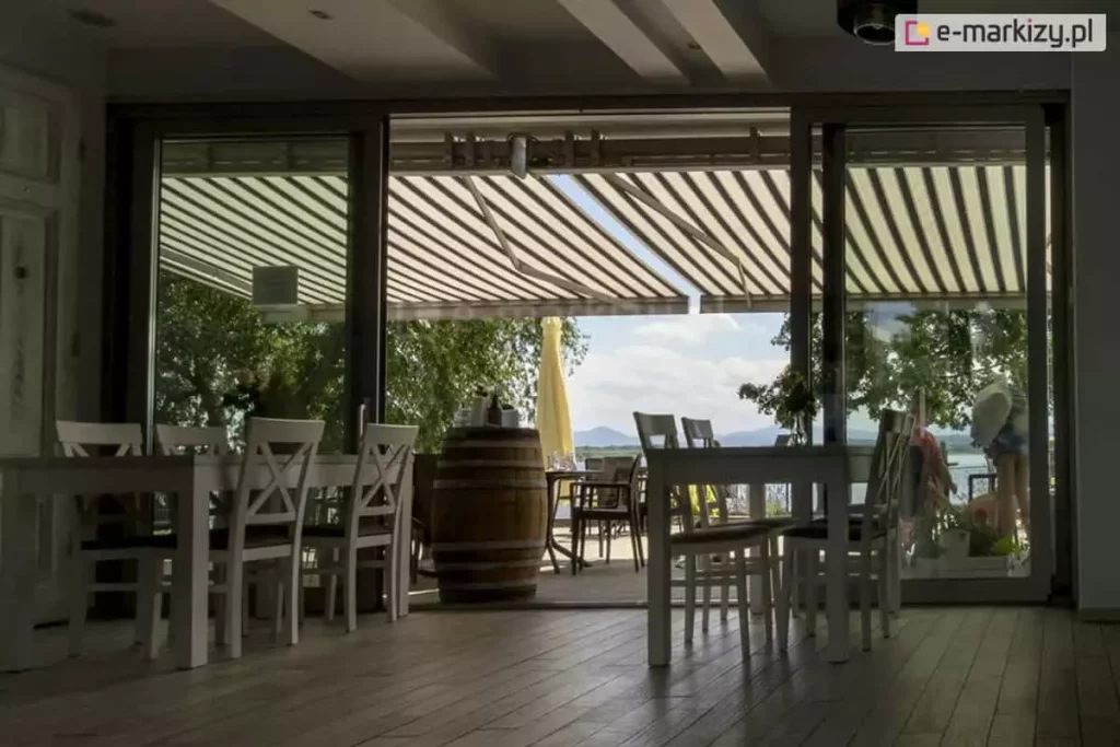 Zdjęcie przedstawia widok z wnętrza restauracji lub kawiarni na zewnętrzny taras, nad którym rozłożona jest duża markiza Corsica firmy Aluprof. Poszycie markizy wykonane jest z tkaniny w szerokie, pionowe pasy w kolorze beżowym i ciemnobrązowym, co nadaje przestrzeni klasyczny i przytulny charakter. Markiza jest w pełni wysunięta, chroniąc gości przed słońcem i tworząc komfortową strefę cienia. Wnętrze lokalu oddzielone jest od tarasu dużymi, przesuwnymi przeszklonymi drzwiami, przez które wpada naturalne światło. Wewnątrz widoczne są białe drewniane stoliki oraz krzesła z krzyżowymi oparciami, typowe dla stylu rustykalnego lub prowansalskiego. Na tarasie, poza zasięgiem głównego cienia markizy, stoi duża drewniana beczka pełniącą funkcję stolika lub dekoracji, a w tle widać parasol ogrodowy oraz sylwetki gór i jeziora pod jasnym, lekko zachmurzonym niebem. Całość kompozycji podkreśla funkcjonalność markizy w obiektach komercyjnych, zapewniając ochronę przeciwsłoneczną przy jednoczesnym zachowaniu estetycznego wyglądu miejsca wypoczynku.