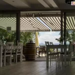 Zdjęcie przedstawia widok z wnętrza restauracji lub kawiarni na zewnętrzny taras, nad którym rozłożona jest duża markiza Corsica firmy Aluprof. Poszycie markizy wykonane jest z tkaniny w szerokie, pionowe pasy w kolorze beżowym i ciemnobrązowym, co nadaje przestrzeni klasyczny i przytulny charakter. Markiza jest w pełni wysunięta, chroniąc gości przed słońcem i tworząc komfortową strefę cienia. Wnętrze lokalu oddzielone jest od tarasu dużymi, przesuwnymi przeszklonymi drzwiami, przez które wpada naturalne światło. Wewnątrz widoczne są białe drewniane stoliki oraz krzesła z krzyżowymi oparciami, typowe dla stylu rustykalnego lub prowansalskiego. Na tarasie, poza zasięgiem głównego cienia markizy, stoi duża drewniana beczka pełniącą funkcję stolika lub dekoracji, a w tle widać parasol ogrodowy oraz sylwetki gór i jeziora pod jasnym, lekko zachmurzonym niebem. Całość kompozycji podkreśla funkcjonalność markizy w obiektach komercyjnych, zapewniając ochronę przeciwsłoneczną przy jednoczesnym zachowaniu estetycznego wyglądu miejsca wypoczynku.