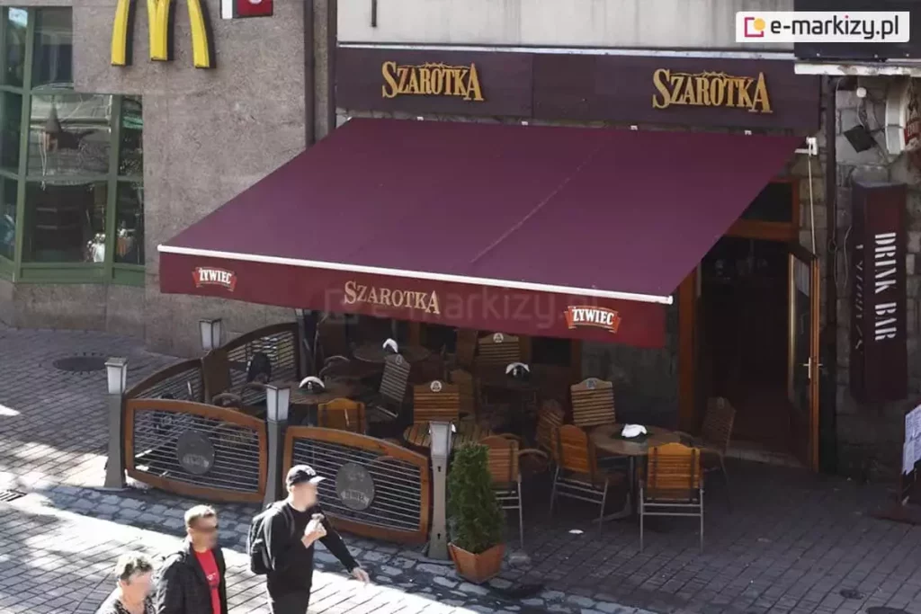 Markiza Jamaica Aluprof z poszyciem w kolorze czerwonego wina oraz nadrukiem reklamowym restauracji "Szarotka" na prostej falbanie zamocowana do ściany nad wejściem do restauracji, zapewnia cień kilku ustawionym pod nią stolikom z krzesłami, tworzącymi przestrzeń dla klientów przy deptaku.