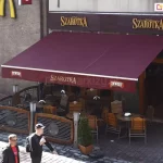 Markiza Jamaica Aluprof z poszyciem w kolorze czerwonego wina oraz nadrukiem reklamowym restauracji "Szarotka" na prostej falbanie zamocowana do ściany nad wejściem do restauracji, zapewnia cień kilku ustawionym pod nią stolikom z krzesłami, tworzącymi przestrzeń dla klientów przy deptaku.