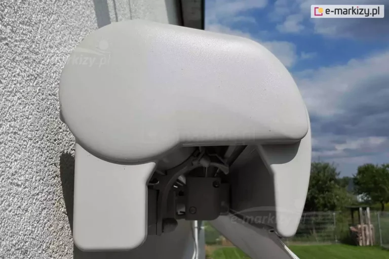 Markiza Palladio Selt w całości złożona widoczna od lewego boku. Pod plastikową zaślepką boczną widoczny wspornik kasety oraz złożone ramię tuż przy belce przedniej, w której widoczny skrawek powiewającej na wietrze falbany. Markiza zamocowana na białej ścianie, obok niej widoczny ogród oraz drzewa z zielonymi liśćmi i zachmurzone niebo.