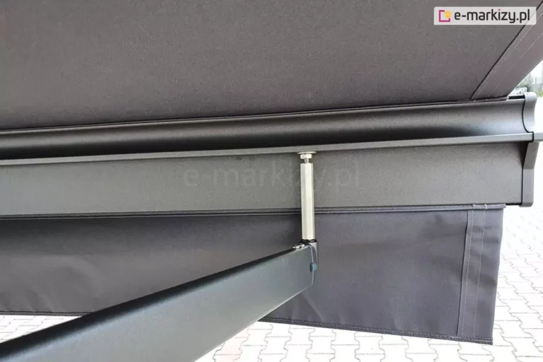 Zbliżenie antracytowej belki przedniej markizy Palladio Selt o charakterystycznym kształcie, z umocowaną w niej prostą falbaną oraz poszyciem w kolorze grafitowym, a także przedłużonym trzpieniem mocowania obniżonego ramienia krzyżowego. W tle widoczna kostka brukowa.