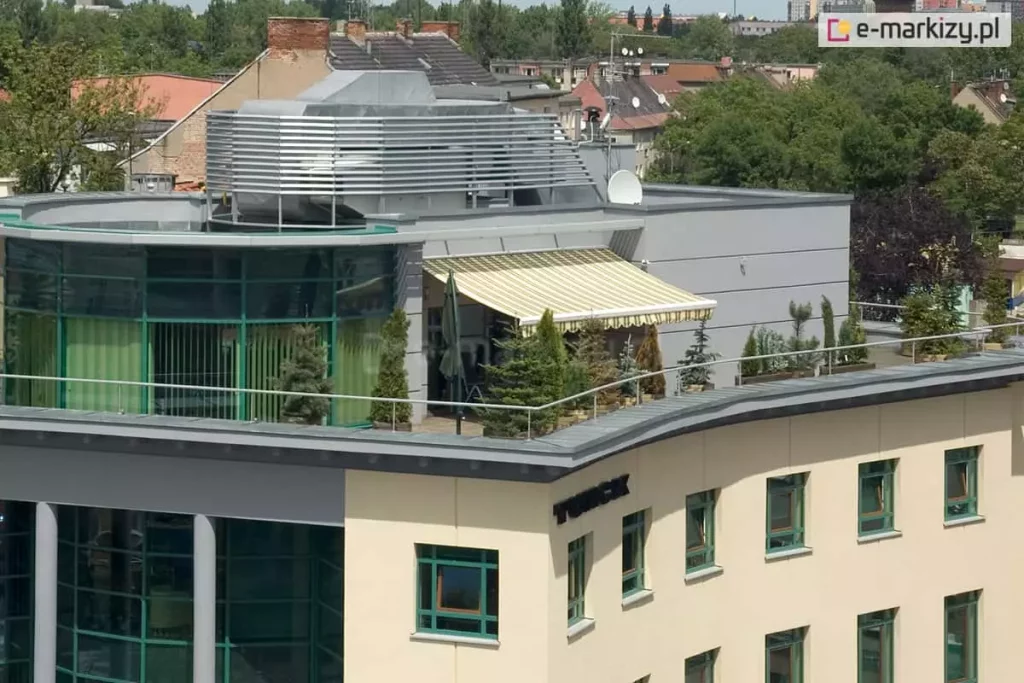 Widok z lotu ptaka na rozległy taras umieszczony na dachu nowoczesnego budynku biurowego lub apartamentowca. Na tarasie rozłożona jest duża osłona przeciwsłoneczna - markiza Palladio Aluprof o poszyciu w jasne, żółto-białe pionowe pasy, wykończona dekoracyjną falbaną na krawędzi. Konstrukcja osłania strefę wypoczynkową, w której ustawione są liczne rośliny iglaste w donicach, tworzące zielony szpaler wzdłuż metalowej barierki zabezpieczającej krawędź dachu. Budynek posiada elewację w kolorze piaskowym z licznymi oknami w zielonych ramach oraz nowoczesną, przeszkloną nadbudówkę o półkolistym kształcie. Na dachu widoczne są również elementy infrastruktury technicznej, w tym srebrne osłony wentylacyjne o lamelowej budowie oraz antena satelitarna. W tle rozpościera się panorama miasta z widocznymi dachami kamienic, zielonymi koronami drzew oraz jasnym, lekko zamglonym niebem.