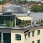 Widok z lotu ptaka na rozległy taras umieszczony na dachu nowoczesnego budynku biurowego lub apartamentowca. Na tarasie rozłożona jest duża osłona przeciwsłoneczna - markiza Palladio Aluprof o poszyciu w jasne, żółto-białe pionowe pasy, wykończona dekoracyjną falbaną na krawędzi. Konstrukcja osłania strefę wypoczynkową, w której ustawione są liczne rośliny iglaste w donicach, tworzące zielony szpaler wzdłuż metalowej barierki zabezpieczającej krawędź dachu. Budynek posiada elewację w kolorze piaskowym z licznymi oknami w zielonych ramach oraz nowoczesną, przeszkloną nadbudówkę o półkolistym kształcie. Na dachu widoczne są również elementy infrastruktury technicznej, w tym srebrne osłony wentylacyjne o lamelowej budowie oraz antena satelitarna. W tle rozpościera się panorama miasta z widocznymi dachami kamienic, zielonymi koronami drzew oraz jasnym, lekko zamglonym niebem.