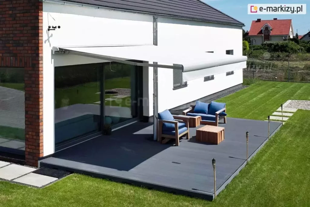 Widok z lotu ptaka na nowoczesny, ciemnoszary taras, nad którym rozłożona jest biała osłona przeciwsłoneczna w postaci markizy tarasowej palladio Aluprof. Konstrukcja posiada ramion podtrzymujące przedni profil, co stabilizuje poszycie zamontowane do białej elewacji budynku. Konstrukcja markizy wykonana została z aluminium malowanego proszkowo na biało i uzupełnionego białą akrylową tkaniną poszycia. Na tarasie znajduje się zestaw mebli ogrodowych wykonanych z jasnego drewna z niebieskimi poduszkami, w tym dwuosobowa sofa, fotel oraz pasujący stolik kawowy. Krawędź tarasu zdobią wbite w ziemię pochodnie bambusowe, a wokół rozciąga się równo przystrzyżony, zielony trawnik z ułożoną ścieżką z jasnych płyt chodnikowych. Budynek posiada duże przeszklenia drzwi tarasowych, w których odbija się otoczenie, oraz fragmenty ściany z czerwonej cegły. W oddali widać sąsiednią posesję z domem o czerwonym dachu, oddzieloną ogrodzeniem z siatki. Całość sceny jest oświetlona jasnym światłem dziennym w pogodny dzień.