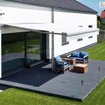 Widok z lotu ptaka na nowoczesny, ciemnoszary taras, nad którym rozłożona jest biała osłona przeciwsłoneczna w postaci markizy tarasowej palladio Aluprof. Konstrukcja posiada ramion podtrzymujące przedni profil, co stabilizuje poszycie zamontowane do białej elewacji budynku. Konstrukcja markizy wykonana została z aluminium malowanego proszkowo na biało i uzupełnionego białą akrylową tkaniną poszycia. Na tarasie znajduje się zestaw mebli ogrodowych wykonanych z jasnego drewna z niebieskimi poduszkami, w tym dwuosobowa sofa, fotel oraz pasujący stolik kawowy. Krawędź tarasu zdobią wbite w ziemię pochodnie bambusowe, a wokół rozciąga się równo przystrzyżony, zielony trawnik z ułożoną ścieżką z jasnych płyt chodnikowych. Budynek posiada duże przeszklenia drzwi tarasowych, w których odbija się otoczenie, oraz fragmenty ściany z czerwonej cegły. W oddali widać sąsiednią posesję z domem o czerwonym dachu, oddzieloną ogrodzeniem z siatki. Całość sceny jest oświetlona jasnym światłem dziennym w pogodny dzień.