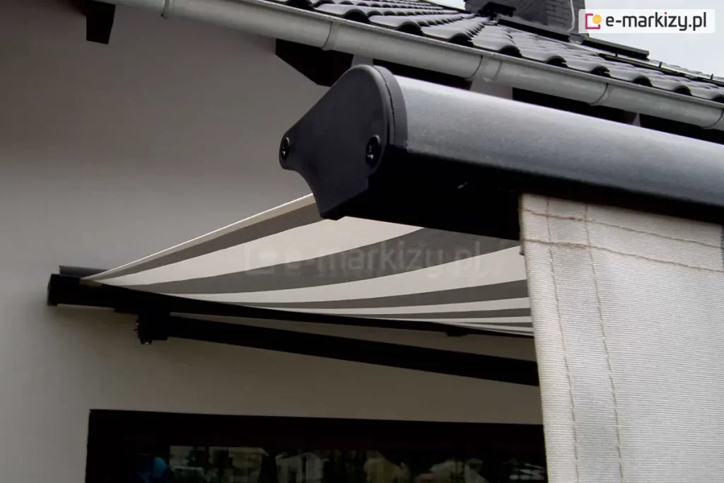 Zdjęcie prezentuje zbliżenie na profil czołowy oraz detale wykończenia markizy Adagio zamontowanej na jasnej elewacji budynku. W pierwszym planie widoczny jest boczny element konstrukcyjny profilu przedniego, wykonany z metalu o grafitowym, matowym wykończeniu, zakończony czarną, plastikową zaślepką z widocznymi śrubami montażowymi. Z profilu czołowego zwiesza się pionowo fragment falbany lub materiału w kolorze kremowym z delikatnym przeszyciem. W dalszej części kadru widać rozpięte poszycie markizy Adagio, które charakteryzuje się nowoczesnym wzorem w szerokie, poziome pasy w kolorach białym i szarym. Tkanina jest mocno napięta, tworząc płaszczyznę chroniącą przed słońcem. Nad konstrukcją markizy Adagio znajduje się fragment dachu pokrytego ciemną dachówką ceramiczną oraz biała rynna dachowa przebiegająca równolegle do krawędzi dachu. Poniżej rozłożonego materiału zarysowuje się ciemna rama okienna lub drzwiowa. Całość kompozycji podkreśla techniczne aspekty wykończenia oraz estetykę dopasowania kolorystycznego markizy Adagio do nowoczesnej architektury domu.