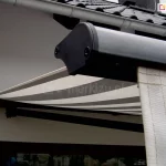 Zdjęcie prezentuje zbliżenie na profil czołowy oraz detale wykończenia markizy Adagio zamontowanej na jasnej elewacji budynku. W pierwszym planie widoczny jest boczny element konstrukcyjny profilu przedniego, wykonany z metalu o grafitowym, matowym wykończeniu, zakończony czarną, plastikową zaślepką z widocznymi śrubami montażowymi. Z profilu czołowego zwiesza się pionowo fragment falbany lub materiału w kolorze kremowym z delikatnym przeszyciem. W dalszej części kadru widać rozpięte poszycie markizy Adagio, które charakteryzuje się nowoczesnym wzorem w szerokie, poziome pasy w kolorach białym i szarym. Tkanina jest mocno napięta, tworząc płaszczyznę chroniącą przed słońcem. Nad konstrukcją markizy Adagio znajduje się fragment dachu pokrytego ciemną dachówką ceramiczną oraz biała rynna dachowa przebiegająca równolegle do krawędzi dachu. Poniżej rozłożonego materiału zarysowuje się ciemna rama okienna lub drzwiowa. Całość kompozycji podkreśla techniczne aspekty wykończenia oraz estetykę dopasowania kolorystycznego markizy Adagio do nowoczesnej architektury domu.