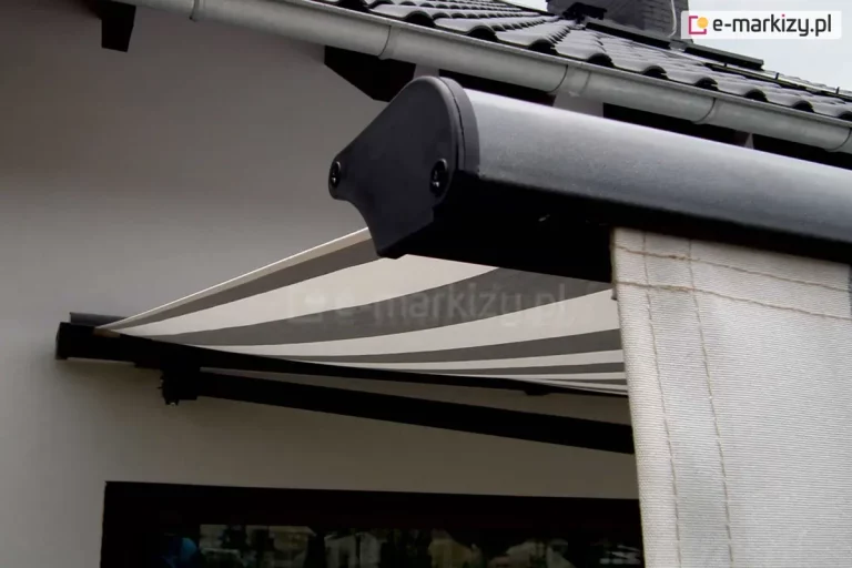 Zdjęcie prezentuje zbliżenie na profil czołowy oraz detale wykończenia markizy Adagio zamontowanej na jasnej elewacji budynku. W pierwszym planie widoczny jest boczny element konstrukcyjny profilu przedniego, wykonany z metalu o grafitowym, matowym wykończeniu, zakończony czarną, plastikową zaślepką z widocznymi śrubami montażowymi. Z profilu czołowego zwiesza się pionowo fragment falbany lub materiału w kolorze kremowym z delikatnym przeszyciem. W dalszej części kadru widać rozpięte poszycie markizy Adagio, które charakteryzuje się nowoczesnym wzorem w szerokie, poziome pasy w kolorach białym i szarym. Tkanina jest mocno napięta, tworząc płaszczyznę chroniącą przed słońcem. Nad konstrukcją markizy Adagio znajduje się fragment dachu pokrytego ciemną dachówką ceramiczną oraz biała rynna dachowa przebiegająca równolegle do krawędzi dachu. Poniżej rozłożonego materiału zarysowuje się ciemna rama okienna lub drzwiowa. Całość kompozycji podkreśla techniczne aspekty wykończenia oraz estetykę dopasowania kolorystycznego markizy Adagio do nowoczesnej architektury domu.