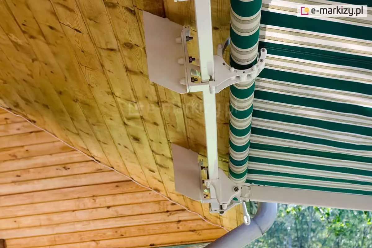 Spadzisty dach z drewnianą podbitką, pod którą ukryto częściowo uchwyty markizy Australia Selt zamocowane do krokwi. Biała konstrukcja współgra kolorystycznie z jasnym drewnem, sprawiając, że markiza staje się integralną częścią budynku. Poszycie w zielono-białe pasy harmonizuje z otoczeniem.