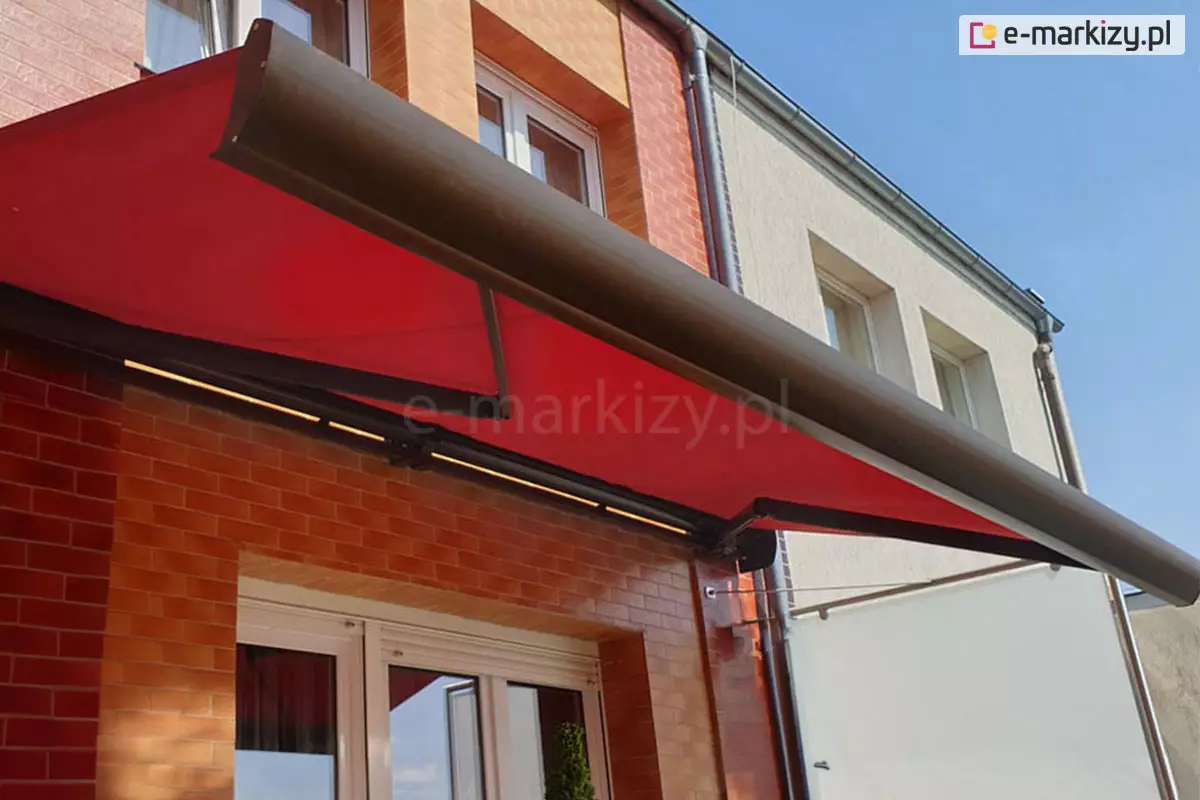 Zdjęcie prezentuje zbliżenie na markizę Corsica Selt zamontowaną na elewacji nowoczesnego budynku wielorodzinnego lub szeregowego. Markiza wyróżnia się intensywnym, czerwonym kolorem poszycia, które mimo, że jest tylko częściowo rozłożone pozostaje idealnie napięte na czarnej konstrukcji ramion. Pod kasetą markizy widoczna jest zintegrowana listwa z oświetleniem LED, która rzuca ciepłe światło na ścianę budynku. Elewacja w miejscu montażu wykonana jest z estetycznych płytek klinkierowych w odcieniach czerwieni i pomarańczu, układanych w poziomie. Produkt osłania duże, białe okna balkonowe. Po prawej stronie widoczny jest fragment sąsiedniego segmentu budynku o jasnej, tynkowanej elewacji, oddzielony pionową rurą spustową. W dolnej części, za barierką, dostrzec można również fragment białej rolety lub przesłony bocznej. Zdjęcie zostało wykonane z perspektywy żabiej, patrząc od dołu ku górze, co podkreśla dynamiczną formę markizy na tle bezchmurnego, błękitnego nieba.