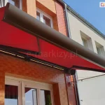 Na ścianie z cegły klinkierowej z oknem o białych ramach i roletach zewnętrznych zamocowano markizę Corsica z brązową konstrukcją i czerwoną tkaniną poszycia oraz zintegrowanym oświetleniem LED w kasecie