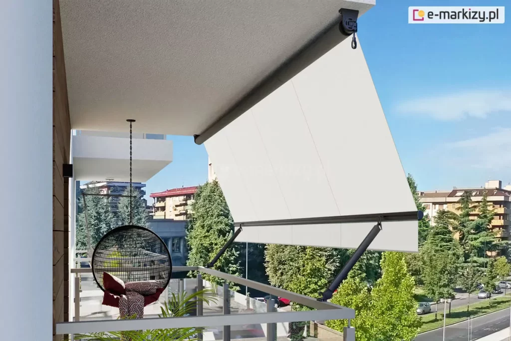 Widok z boku na nowoczesny balkon wyposażony w markizę balkonową z ramionami poręczowymi, zamontowaną pod sufitem wnęki. Markiza posiada poszycie z gładkiej tkaniny w kolorze kremowym lub bardzo jasnym beżu, która jest rozwinięta i napięta pod kątem, wychodząc poza obrys barierki. Konstrukcja wspiera się na dwóch ciemnych, antracytowych ramionach bocznych przymocowanych do balustrady, które utrzymują dolną belkę dociążającą. Na suficie widoczna jest rura nawojowa z mechanizmem korbowym zakończonym oczkiem. Na tarasie, w cieniu osłony, znajduje się wiszący fotel typu kokon o czarnym, ażurowym splotcie, zawieszony na metalowym łańcuchu, wyłożony wzorzystymi poduszkami. Obok fotela widać fragmenty zielonych roślin doniczkowych. Balustrada balkonu jest minimalistyczna, wykonana z jasnych profili metalowych z wypełnieniem szklanym. W tle rozpościera się widok na miejską panoramę z wielopiętrowymi budynkami mieszkalnymi, gęstą zielenią drzew oraz fragmentem ulicy z zaparkowanymi samochodami pod bezchmurnym, błękitnym niebem.