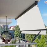 Widok z boku na nowoczesny balkon wyposażony w markizę balkonową z ramionami poręczowymi, zamontowaną pod sufitem wnęki. Markiza posiada poszycie z gładkiej tkaniny w kolorze kremowym lub bardzo jasnym beżu, która jest rozwinięta i napięta pod kątem, wychodząc poza obrys barierki. Konstrukcja wspiera się na dwóch ciemnych, antracytowych ramionach bocznych przymocowanych do balustrady, które utrzymują dolną belkę dociążającą. Na suficie widoczna jest rura nawojowa z mechanizmem korbowym zakończonym oczkiem. Na tarasie, w cieniu osłony, znajduje się wiszący fotel typu kokon o czarnym, ażurowym splotcie, zawieszony na metalowym łańcuchu, wyłożony wzorzystymi poduszkami. Obok fotela widać fragmenty zielonych roślin doniczkowych. Balustrada balkonu jest minimalistyczna, wykonana z jasnych profili metalowych z wypełnieniem szklanym. W tle rozpościera się widok na miejską panoramę z wielopiętrowymi budynkami mieszkalnymi, gęstą zielenią drzew oraz fragmentem ulicy z zaparkowanymi samochodami pod bezchmurnym, błękitnym niebem.
