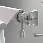 Zbliżenie techniczne na system mocowania markizy balkonowej Italia, prezentujące uniwersalne uchwyty ścienno-sufitowe. Na zdjęciu widoczna jest boczna głowica markizy w kolorze jasnoszarym, zamontowana w odlewanym, aluminiowym uchwycie o budowie modułowej. Konstrukcja uchwytu posiada prostopadłe płaszczyzny z otworami, co pozwala na jego przykręcenie zarówno do pionowej ściany, jak i poziomego sufitu. W centralnej części detalu widoczna jest przekładnia z wyciągniętym pionowo w dół plastikowym uchem, służącym do obsługi za pomocą korby. Belka nawojowa z nawiniętą jasną tkaniną jest stabilnie osadzona w gnieździe uchwytu. Po prawej stronie u dołu znajduje się szary prostokąt z białym napisem informacyjnym, od którego zielona linia prowadzi do punktu styku głowicy z uchwytem, podkreślając precyzję dopasowania elementów. W tle widoczna jest również przednia belka obciążająca z zaokrągloną zaślepką boczną.