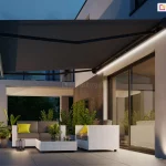 Przestrony taras przy apartamencie, na którym ustawiono komplet mebli wypoczynkowych z technorattanu w kolorze białym z brązowymi poduchami i żółtymi dodatkami oraz rośliny doniczkowe, oświetla wieczorem taśma led w kasecie markizy Corsica rozwiniętej nad meblami