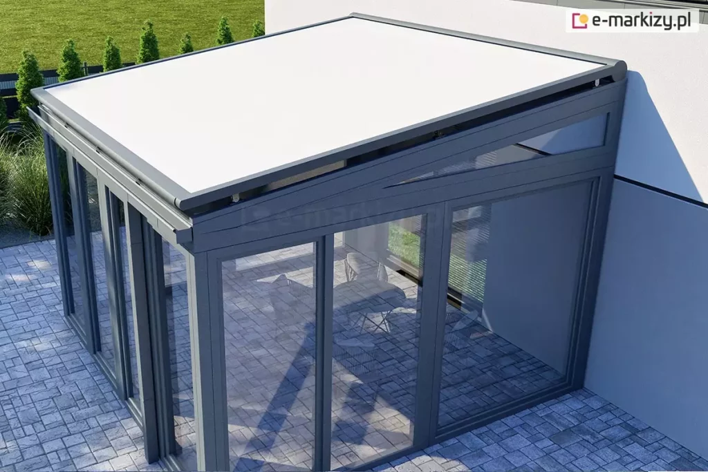 nowoczesny ogród zimowy o konstrukcji aluminiowo-szklanej, widziany z góry pod lekkim kątem, od strony zewnętrznego narożnika. Na szklanym dachu obiektu zainstalowano zewnętrzny system Veranda V-Ziiip aluprof z jasnym, białym poszyciem, które jest w pełni rozwinięte i precyzyjnie napięte. Konstrukcja nośna ogrodu zimowego, w tym ramy okienne, profile dachowe oraz prowadnice verandy V-Ziiip, są polakierowane na elegancki, matowy kolor antracytowy. Ściany boczne i frontowe obiektu są w całości przeszklone, co pozwala dostrzec wnętrze z jasną, brukowaną posadzką oraz zarysem mebli wypoczynkowych. Ogród zimowy przylega do jasnej elewacji budynku, a wokół niego rozciąga się starannie utrzymany trawnik oraz rząd tui wzdłuż ogrodzenia w tle. Perspektywa zdjęcia uwypukla techniczne aspekty montażu osłony przeciwsłonecznej oraz nowoczesny design całej bryły architektonicznej, łączącej ciemne profile z jasnym materiałem markizy.