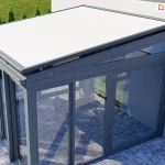 nowoczesny ogród zimowy o konstrukcji aluminiowo-szklanej, widziany z góry pod lekkim kątem, od strony zewnętrznego narożnika. Na szklanym dachu obiektu zainstalowano zewnętrzny system Veranda V-Ziiip aluprof z jasnym, białym poszyciem, które jest w pełni rozwinięte i precyzyjnie napięte. Konstrukcja nośna ogrodu zimowego, w tym ramy okienne, profile dachowe oraz prowadnice verandy V-Ziiip, są polakierowane na elegancki, matowy kolor antracytowy. Ściany boczne i frontowe obiektu są w całości przeszklone, co pozwala dostrzec wnętrze z jasną, brukowaną posadzką oraz zarysem mebli wypoczynkowych. Ogród zimowy przylega do jasnej elewacji budynku, a wokół niego rozciąga się starannie utrzymany trawnik oraz rząd tui wzdłuż ogrodzenia w tle. Perspektywa zdjęcia uwypukla techniczne aspekty montażu osłony przeciwsłonecznej oraz nowoczesny design całej bryły architektonicznej, łączącej ciemne profile z jasnym materiałem markizy.