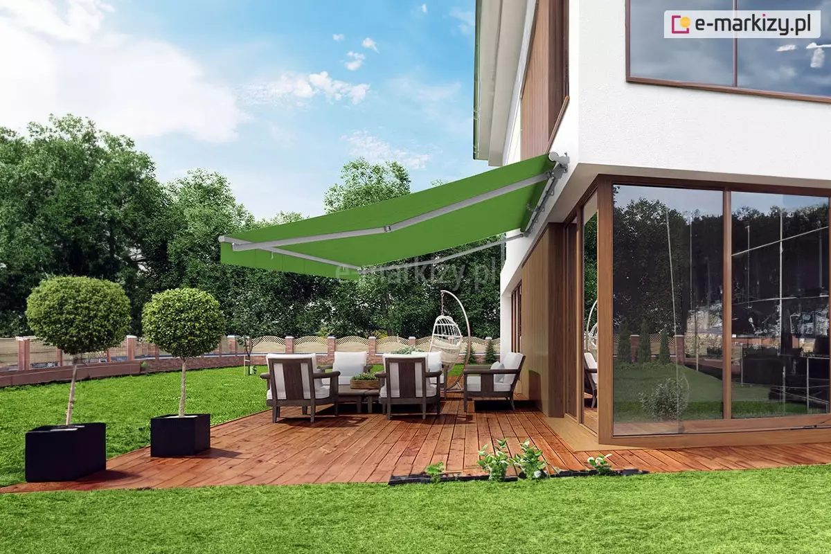 Zdjęcie prezentuje nowoczesną wizualizację tarasu przy dwukondygnacyjnym budynku, na którym zainstalowano dużą markizę tarasową w intensywnym, zielonym kolorze. Markiza ma konstrukcję w kolorze jasnoszarym (srebrnym) i jest zamontowana do białej elewacji budynku, chroniąc przed słońcem strefę wypoczynkową. Pod osłoną znajduje się drewniany taras wykonany z desek w ciepłym odcieniu brązu, na którym ustawiono komplet mebli ogrodowych: cztery fotele z ciemną konstrukcją i białymi poduszkami oraz niski stolik kawowy. W tle, obok mebli, widoczne jest wiszące krzesło typu „kokon” na stabilnym stelażu. Parter budynku charakteryzuje się dużymi, narożnymi przeszkleniami w brązowych ramach, które odbijają otaczającą zieleń. Ogród jest starannie utrzymany, z idealnie przyciętym trawnikiem, na którym ustawiono dwie nowoczesne, czarne, wysokie donice z formowanymi krzewami o kulistym kształcie. Posesja jest ogrodzona płotem o dekoracyjnym wzorze, a za nim widoczna jest gęsta ściana wysokich, liściastych drzew pod jasnobłękitnym niebem z rozproszonymi chmurami.