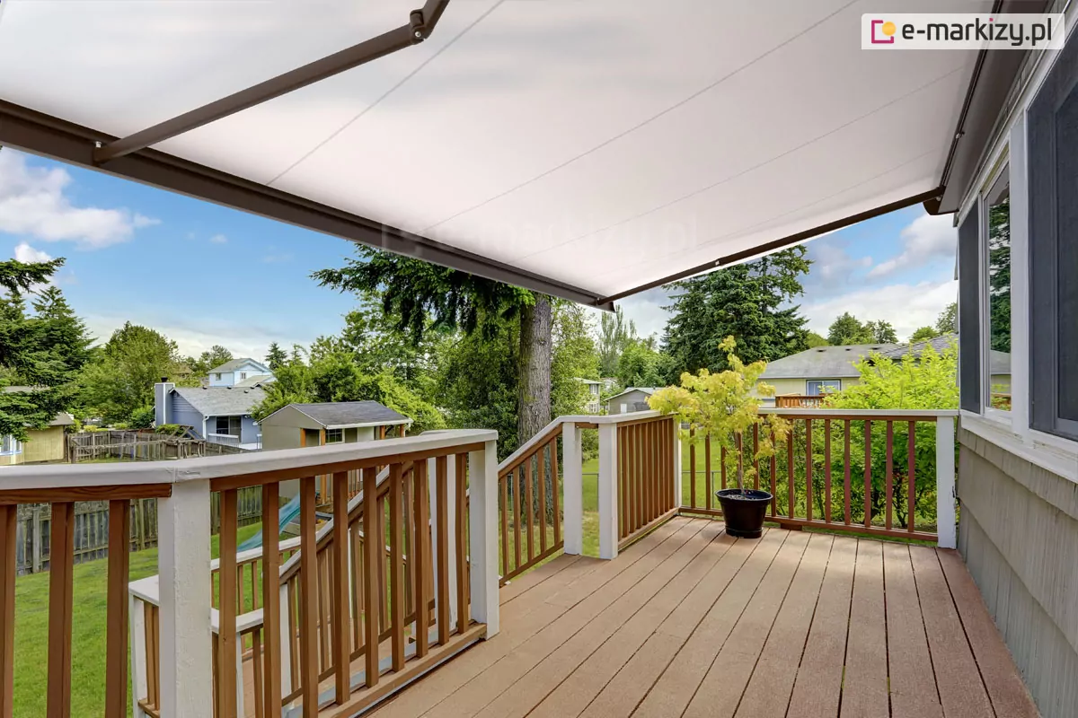 Zdjęcie prezentuje perspektywę z poziomu desek tarasowych na przestronny, drewniany taras osłonięty w całości przez rozłożoną markizę Corsica. Poszycie markizy jest wykonane z jasnej, niemal białej tkaniny, która przepuszcza łagodne, rozproszone światło, tworząc komfortową strefę wypoczynku. Konstrukcja wspierająca materiał, w tym widoczne po lewej stronie ramię przegubowe, wykończona jest w ciemnym, brązowym lub antracytowym kolorze, kontrastującym z jasnym materiałem. Taras posiada balustradę wykonaną z pionowych, drewnianych szczebli osadzonych między solidnymi, białymi słupkami, a jego podłogę tworzą deski kompozytowe w naturalnym odcieniu drewna. W narożniku tarasu ustawiona jest ciemna donica z ozdobnym, jasnozielonym krzewem. Z tarasu rozpościera się widok na osiedle domów jednorodzinnych z zadbanymi trawnikami, drzewami iglastymi oraz elementami architektury ogrodowej, takimi jak domek narzędziowy ze zjeżdżalnią. Niebo jest częściowo zachmurzone, z wyraźnymi prześwitami błękitu, co w połączeniu z markizą Corsica sugeruje idealne warunki do spędzania czasu na zewnątrz w słoneczny dzień.
