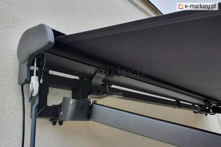 Widok lewej strony markizy Palladio zawieszonej na ścianie z białą elewacją, konstrukcja markizy w kolorze srebrzysty grafit z widocznym mocowaniem kasety do belki kwadrat oraz umocowaniem ramienia krzyżowego obniżonego, ponad którym widać rozwinięte poszycie markizy z tkaniny akrylowej w kolorze Antracita. Po lewej stronie markizy widoczna zaślepka boczna z pcv, pod którą wystaje przewód zasilający silnika markizy oraz zaczep przekładni do awaryjnego otwierania markizy za pomocą korby.