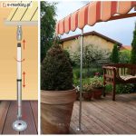 Po lewej prosty schemat teleskopowego słupka montażowego do markizy z strzałkami pokazującymi ruchy, po prawej zdjęcie metalowego słupka podtrzymującego pomarańczowo-białą, falującą markizę nad drewnianym tarasem z donicami z roślinami i drewnianą ławką, w tle ogród i dom.