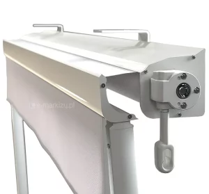 Zbliżenie techniczne na mechanizm i kasetę markizy balkonowej w kolorze białym. Zdjęcie prezentuje profil aluminiowej kasety z widocznym systemem sterowania ręcznego w postaci przekładni z uchem na korbę. Na górnej części kasety znajdują się dwa stalowe uchwyty montażowe typu sufitowego. Z kasety wysunięta jest belka frontowa z fragmentem białego poszycia, co obrazuje sposób chowania się tkaniny i domykania profilu. Konstrukcja jest malowana proszkowo na wysoki połysk, a widoczne śruby montażowe podkreślają techniczny charakter produktu.