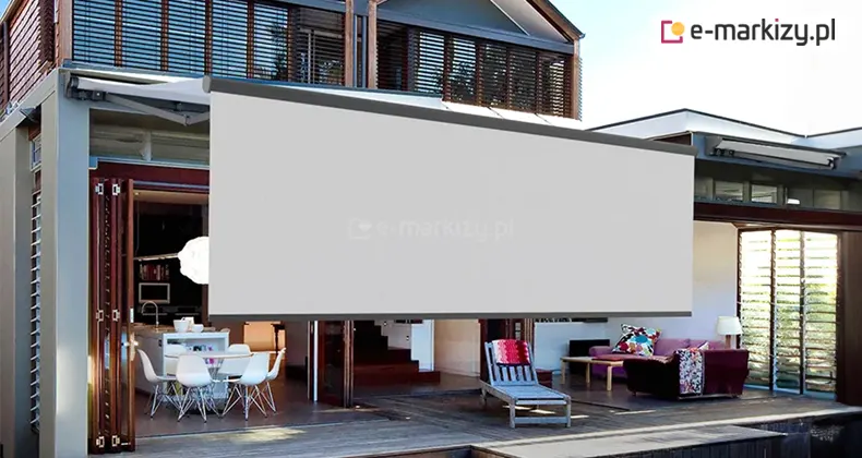 Markiza Jamaica Selt z falbaną rozwijaną zamocowana na ścianie piętrowego domku nad tarasem. Ponad markiza widoczne okna piętra osłonięte żaluzjami fasadowymi. Na tarasie ustawiono leżak do opalania, a zewnętrzna ściana została przesunięta na lewo otwierając przestrzeń wewnętrzną domu, w której widać po prawej kanapę ze stolikiem i lampką, a po lewej stół z krzesłami w białym kolorze. Długa falbana skrywa pozostałą część wnętrza.