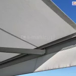 Rozłożona markiza tarasowa Palladio Aluprof widoczna w zbliżeniu od dołu pod kątem, na tle bezchmurnego, błękitnego nieba. Poszycie markizy wykonane jest z gładkiej tkaniny w jasnoszarym, niemal białym kolorze. Materiał jest napięty na konstrukcji, a na jego powierzchni widoczne są podłużne, regularne przeszycia łączące poszczególne bryty tkaniny. Od czoła markizy, u dołu przedniej belki, zwiesza się prosta, klasyczna falbana o prostokątnym kroju, wykończona na dolnej krawędzi lamówką w pasującym, szarym odcieniu. Konstrukcja nośna oraz profile aluminiowe, w tym widoczne przegubowe ramię mechaniczne podtrzymujące poszycie, są lakierowane proszkowo na jednolity, jasnoszary matowy kolor, idealnie współgrający z barwą materiału. Ramię markizy jest częściowo złożone, stabilizując wysunięte poszycie. Aluminiowa belka przednia zamykająca kasetę ma nowoczesny, zaokrąglony kształt i jest wykończona z boku estetyczną, dopasowaną zaślepką z tworzywa sztucznego. Światło słoneczne pada na markizę z boku, tworząc delikatne cienie pod konstrukcją ramienia oraz na powierzchni falbany, co podkreśla przestrzenną strukturę i techniczną budowę systemu osłonowego.