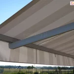 Widok łączenia ramienia markizy Jamaica Aluprof z jej belką przednią, w której mocowane jest poszycie oraz prosta falbana. Jasna konstrukcja oraz poszycie w kremowe i beżowe pasy osłaniają przestrzeń znajdującą się pod osłoną. Nad poszyciem markizy widoczne niebieskie niebo, pod poszyciem widoczna okolica, pełna łąk i płotów oraz sąsiednie domki.
