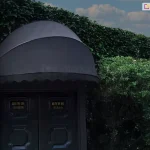 Na zdjęciu widoczna markiza koszowa półokrągła zamontowana nad wejściem do budynku. Poszycie markizy wykonane jest z gładkiego materiału w bardzo ciemnym, niemal czarnym lub głębokim grafitowym kolorze, który nadaje instalacji elegancki i prestiżowy charakter. Dolna krawędź osłony jest wykończona ozdobną, regularnie pofalowaną falbaną. Markiza osłania masywne, dwuskrzydłowe drzwi wykonane z ciemnego drewna lub metalu, ozdobione klasycznymi płycinami oraz detalami w kolorze złotym, które mogą być tabliczkami informacyjnymi lub elementami dekoracyjnymi. Całość konstrukcji jest wkomponowana w gęstą, starannie utrzymaną roślinność ogrodową. Nad markizą oraz po jej bokach rozpościera się ściana zieleni z pnączy i krzewów o drobnych liściach, tworząc naturalne, cieniste obramowanie. W tle, ponad linią roślinności, widoczne jest pogodne niebo z delikatnie zarysowanymi, jasnymi chmurami w świetle późnego popołudnia. Kompozycja zdjęcia podkreśla luksusowy wygląd obiektu oraz funkcjonalną rolę markizy jako elementu chroniącego wejście przed słońcem i deszczem, przy jednoczesnym zachowaniu harmonii z naturalnym otoczeniem.