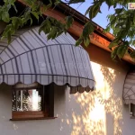 Zdjęcie przedstawia dwie identyczne markizy koszowe o półkolistym kształcie, zamontowane nad oknami na parterze jasnego budynku. Markizy posiadają eleganckie poszycie w stonowanej kolorystyce, składające się z pionowych pasów w różnych odcieniach szarości i beżu, co nadaje im klasyczny wygląd. Konstrukcja każdej osłony opiera się na białym stelażu, a dolne krawędzie są wykończone dekoracyjną, falistą falbaną z ciemniejszą lamówką. Markizy są przytwierdzone do elewacji o tynku w kolorze złamanej bieli, bezpośrednio pod drewnianym podbitką dachu w ciepłym odcieniu brązu. Okna posiadają brązowe ramy, a w ich szybach odbija się zieleń ogrodu. Na elewację padają wyraźne cienie rzucane przez pobliskie drzewa, których liście są widoczne w górnej i bocznej części kadru. Scena jest skąpana w naturalnym, popołudniowym świetle słonecznym, co podkreśla teksturę materiałów i tworzy spokojną, domową atmosferę. Instalacja pełni funkcję zarówno praktyczną, chroniąc wnętrza przed nadmiernym nasłonecznieniem, jak i dekoracyjną, wzbogacając estetykę bryły budynku.