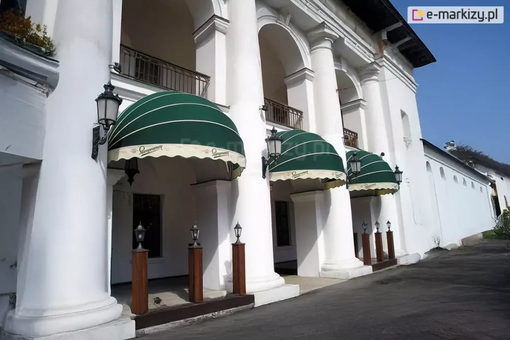 Zdjęcie przedstawia fragment zabytkowej białej budowli z kolumnadą, w której łukach zamontowano trzy eleganckie markizy koszowe o półkolistym kształcie. Poszycie markiz wykonane jest z tkaniny w głębokim, ciemnozielonym kolorze, którą zdobią cienkie, poziome, białe pasy biegnące wzdłuż łuków stelaża. Dolne krawędzie markiz wykończone są prostymi, jasnymi falbanami w kolorze kremowym, na których nadrukowany jest ozdobny, czarny logotyp marki „Staropramen”. Każda z markiz jest precyzyjnie osadzona między masywnymi, białymi kolumnami o klasycznych bazach i kapitelach. Na kolumnach, po bokach markiz, zamocowane są stylizowane czarne latarnie ścienne z przeszklonymi kloszami. Pod zadaszeniami, na podestach, ustawiono mniejsze, stojące lampy ogrodowe na drewnianych słupkach. Budynek posiada liczne arkady i balkony z ozdobnymi, ciemnymi balustradami na wyższej kondygnacji. Przed budynkiem przebiega asfaltowa droga lub plac. Całość scenerii jest oświetlona jasnym, dziennym światłem pod bezchmurnym, błękitnym niebem, co podkreśla kontrast między zielenią osłon a bielą elewacji i nadaje miejscu prestiżowy charakter lokalu gastronomicznego.
