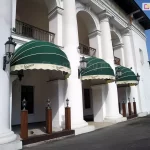 Zdjęcie przedstawia fragment zabytkowej białej budowli z kolumnadą, w której łukach zamontowano trzy eleganckie markizy koszowe o półkolistym kształcie. Poszycie markiz wykonane jest z tkaniny w głębokim, ciemnozielonym kolorze, którą zdobią cienkie, poziome, białe pasy biegnące wzdłuż łuków stelaża. Dolne krawędzie markiz wykończone są prostymi, jasnymi falbanami w kolorze kremowym, na których nadrukowany jest ozdobny, czarny logotyp marki „Staropramen”. Każda z markiz jest precyzyjnie osadzona między masywnymi, białymi kolumnami o klasycznych bazach i kapitelach. Na kolumnach, po bokach markiz, zamocowane są stylizowane czarne latarnie ścienne z przeszklonymi kloszami. Pod zadaszeniami, na podestach, ustawiono mniejsze, stojące lampy ogrodowe na drewnianych słupkach. Budynek posiada liczne arkady i balkony z ozdobnymi, ciemnymi balustradami na wyższej kondygnacji. Przed budynkiem przebiega asfaltowa droga lub plac. Całość scenerii jest oświetlona jasnym, dziennym światłem pod bezchmurnym, błękitnym niebem, co podkreśla kontrast między zielenią osłon a bielą elewacji i nadaje miejscu prestiżowy charakter lokalu gastronomicznego.