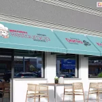 Trzy nowoczesne markizy koszowe o sztywnym, trójkątnym profilu i błękitnym poszyciu w subtelne pionowe prążki, zamontowane w rzędzie na białej elewacji restauracji. Nadruki reklamowe na markizach wykonano w kolorach czerwonym, czarnym i białym: pierwsza od lewej przedstawia logo z kucharzem i napis Restauracja Mała Polinka, druga napis Smaczne Dania Domowe z ikoną serwowania potraw, a trzecia napis Pyszna Kawa oraz Herbatka z grafiką filiżanki. Osłony posiadają prostą falbanę wykończoną białą lamówką i są zamontowane nad dużymi witrynami lokalu gastronomicznego. Pod markizami znajduje się letni ogródek z nowoczesnymi, plecionymi krzesłami i stolikami, a w szybach odbijają się zaparkowane samochody oraz sąsiedni budynek. Powyżej markiz elewacja ozdobiona jest poziomym pasem w kolorze ciemnoszarym z dekoracyjnym gzymsem.