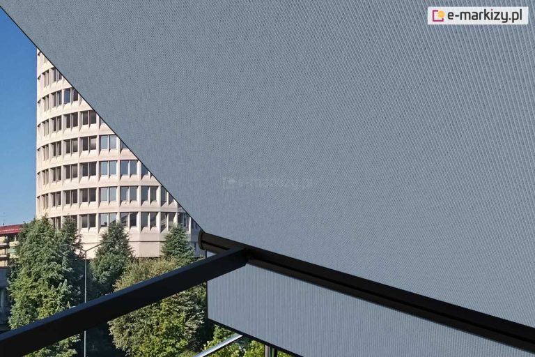 Widok z balkonu lub tarasu na rozwiniętą markizę zewnętrzną Italia wykonaną z tkaniny akrylowej w kolorze jasnoszarym o drobnoziarnistym splotcie. Materiał markizy jest napięty na czarnej, aluminiowej konstrukcji składającej się z poziomej belki dolnej oraz prostopadłych ramion bocznych. W lewej części kadru, za krawędzią poszycia, widoczna jest miejska panorama z charakterystycznym, wysokim budynkiem o cylindrycznym kształcie i jasnej elewacji z gęsto rozmieszczonymi oknami. Poniżej budynku znajdują się korony zielonych drzew liściastych i iglastych, które wypełniają dolną część tła. Całość ujęcia jest oświetlona naturalnym światłem dziennym pod kątem, co uwypukla fakturę materiału markizy oraz tworzy kontrast między zacienioną strefą pod osłoną a jasnym tłem urbanistycznym.