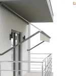 Wizualizacja techniczna markizy balkonowej z ramionami ściennymi, widok z boku. System składa się z aluminiowej kasety w kolorze ciemnoszarym, zamontowanej pod stropem, oraz dwóch sztywnych ramion bocznych przytwierdzonych bezpośrednio do elewacji budynku po obu stronach wnęki balkonowej. Ramiona są nachylone pod kątem, wypychając belkę frontową z jasnokremowym poszyciem poza obrys balkonu. Markiza Aluprof posiada krótką, prostą falbanę. Pod zadaszeniem znajduje się balkon z metalową balustradą oraz przeszklone drzwi balkonowe, obok których zamontowano dwa czarne, pionowe kinkiety elewacyjne. Tło jest białe, co podkreśla detale konstrukcyjne systemu osłonowego.
