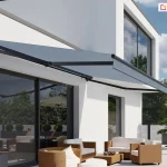 Nowoczesna szara markiza Australia zamontowana na białej, gładkiej elewacji piętrowego budynku mieszkalnego o minimalistycznej architekturze. Poszycie markizy w kolorze grafitowym jest w pełni rozwinięte i napięte na ciemnych, niemal czarnych ramionach przegubowych, rzucając wyraźny cień na taras oraz duże przeszklenia parteru. Konstrukcja markizy Australia charakteryzuje się brakiem pełnej kasety, co eksponuje jej techniczne detale montażowe przytwierdzone powyżej linii okien tarasowych. Pod markizą znajduje się zaaranżowana strefa wypoczynkowa z jasnobrązowymi meblami technorattanowymi z białymi poduszkami, ustawionymi na jasnoszarych płytach tarasowych. W tle widoczny jest ogród z zielonymi drzewami, krzewami formowanymi w kule oraz dodatkowe wyposażenie ogrodowe w postaci beżowego parasola bocznego i drewnianych leżaków o ergonomicznym kształcie. Całość kompozycji przedstawia produkt w pełnym słońcu na tle bezchmurnego, błękitnego nieba, podkreślając funkcję ochronną markizy Australia przed intensywnym promieniowaniem słonecznym.