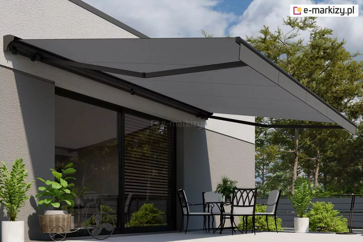 Zdjęcie prezentuje nowoczesną i minimalistyczną aranżację tarasu, nad którym rozłożona została wielkogabarytowa markiza tarasowa Jamaica Selt z opcjonalnym daszkiem. Poszycie markizy oraz cała jej konstrukcja, w tym ramiona przegubowe, profil przedni i daszek, utrzymane są w eleganckiej, jednolitej kolorystyce ciemnego grafitu lub antracytu. Tkanina jest gładka, bez wzorów, co podkreśla surowy i luksusowy charakter osłony. Markiza jest zamontowana do jasnoszarej, tynkowanej elewacji budynku o prostej bryle. Poniżej znajduje się strefa wypoczynkowa wyposażona w stylowe meble ogrodowe. Widoczny jest stół z szklanym blatem oraz cztery metalowe krzesła z ozdobnymi oparciami o geometrycznym wzorze, wyłożone jasnymi poduszkami. Po lewej stronie, przy dużym przesuwnym oknie tarasowym, ustawiono donice z zielonymi roślinami, w tym jedną umieszczoną w dekoracyjnym, wiklinowym koszu na stojaku przypominającym rower. Taras jest wyłożony jasnymi, wielkoformatowymi płytami. W tle widoczne są gęste korony drzew oraz błękitne niebo z delikatnymi chmurami, co sugeruje słoneczny dzień, w którym markiza zapewnia optymalny cień dla użytkowników tarasu.