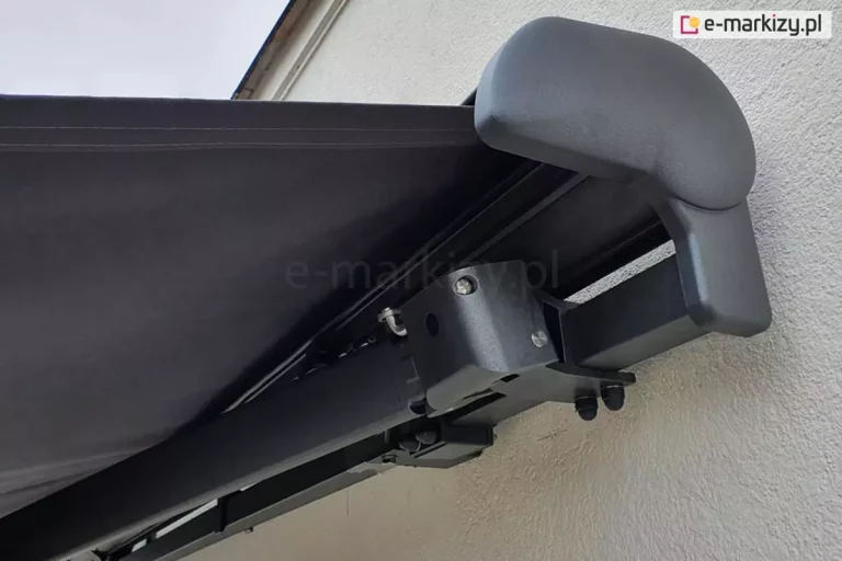 Markiza Palladio Selt zamocowana na ścianie z białym tynkiem strukturalnym, widoczna od prawego boku, konstrukcja w kolorze srebrzysty grafit, tkanina w kolorze antracytowym, widoczna zaślepka boczna z PCV w kolorze konstrukcji oraz mocowanie ramienia na belce kwadratowej.