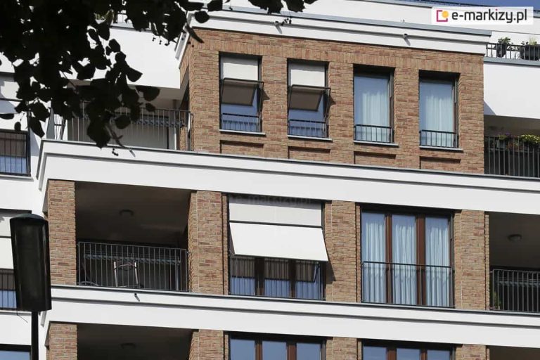 Zdjęcie prezentuje fragment nowoczesnej elewacji budynku wykonanej z jasnobrązowej cegły klinkierowej z białymi, szerokimi pasami międzykondygnacyjnymi. Na fasadzie zainstalowano systemy markizolet zewnętrznych z tkaniną w kolorze kremowym lub bardzo jasnym szarym. W centralnej części widoczne jest duże, dwuskrzydłowe okno z rozwiniętą osłoną, której dolna część została odchylona pod kątem za pomocą bocznych ramion, tworząc daszek osłaniający przeszklenie przed słońcem. Na wyższej kondygnacji znajdują się mniejsze, pojedyncze okna, z których dwa wyposażone są w analogiczne markizolety, również w pozycji uchylonej. Pozostałe okna posiadają jedynie wewnętrzne firany lub są odsłonięte, co ukazuje zróżnicowanie w konfiguracji osłon przez użytkowników. Konstrukcja ramion i prowadnic jest minimalistyczna, w ciemnym kolorze, co kontrastuje z jasnym materiałem osłonowym. Całość kompozycji dopełniają balkony z ażurowymi, ciemnymi balustradami oraz fragment korony drzewa widoczny w lewym górnym rogu.