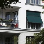 Zdjęcie prezentuje fragment białej, klasycznej elewacji budynku mieszkalnego z loggiami i dużymi przeszkleniami. Głównym elementem jest zainstalowana w szerokiej wnęce okiennej markizoleta w kolorze ciemnozielonym. Osłona jest częściowo rozwinięta, przy czym jej dolna sekcja została odchylona na zewnątrz za pomocą bocznych ramion, tworząc sztywny daszek pod kątem. Górna część tkaniny pozostaje w płaszczyźnie pionowej, przylegając do górnej ramy okna. Po lewej stronie widoczny jest przylegający balkon z ciemną, metalową balustradą, na której zawieszono donice z kwiatami oraz suszarkę z ubraniami. Konstrukcja markizolety, w tym prowadnice i belka dolna, wykonana jest w ciemnym kolorze, kontrastującym z jasną fasadą. Roślinność w postaci liści drzew widoczna na pierwszym planie oraz z boku kadru nadaje zdjęciu naturalny kontekst i sugeruje nasłonecznienie obiektu.