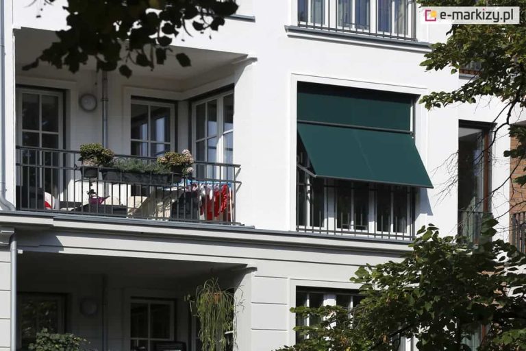 Zdjęcie prezentuje fragment białej, klasycznej elewacji budynku mieszkalnego z loggiami i dużymi przeszkleniami. Głównym elementem jest zainstalowana w szerokiej wnęce okiennej markizoleta w kolorze ciemnozielonym. Osłona jest częściowo rozwinięta, przy czym jej dolna sekcja została odchylona na zewnątrz za pomocą bocznych ramion, tworząc sztywny daszek pod kątem. Górna część tkaniny pozostaje w płaszczyźnie pionowej, przylegając do górnej ramy okna. Po lewej stronie widoczny jest przylegający balkon z ciemną, metalową balustradą, na której zawieszono donice z kwiatami oraz suszarkę z ubraniami. Konstrukcja markizolety, w tym prowadnice i belka dolna, wykonana jest w ciemnym kolorze, kontrastującym z jasną fasadą. Roślinność w postaci liści drzew widoczna na pierwszym planie oraz z boku kadru nadaje zdjęciu naturalny kontekst i sugeruje nasłonecznienie obiektu.