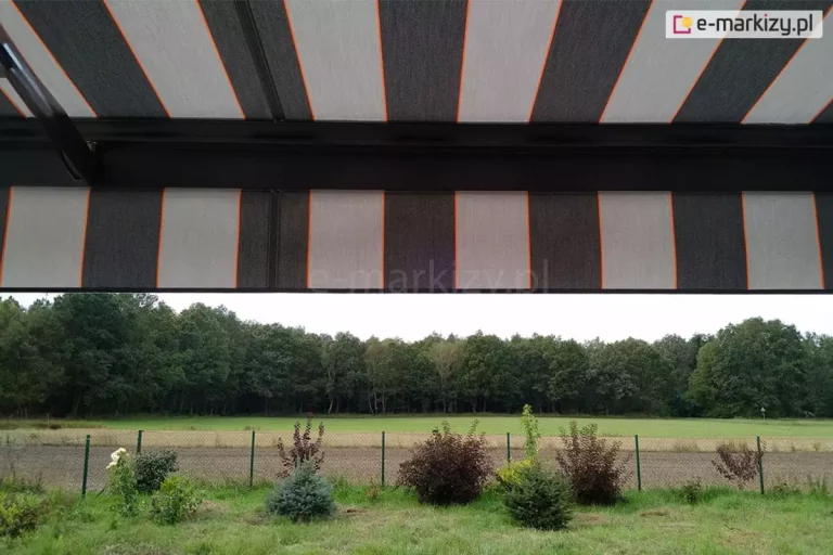 widok z wnętrza tarasu na otaczający krajobraz, częściowo przesłonięty przez nowoczesną markizę tarasową Palladio. Poszycie markizy wykonane z tkaniny akrylowej powlekanej teflonem, charakteryzuje się rytmicznym układem szerokich pasów w kolorze ciemnografitowym oraz jasnoszarym, które są oddzielone od siebie cienkimi, pionowymi liniami w kolorze intensywnej pomarańczy. Materiał jest napięty na czarnej, aluminiowej konstrukcji, której poprzeczna belka czołowa widoczna jest w górnej części kadru. Markiza posiada również prostą falbanę o tym samym wzorze, która zwisa z profilu, ograniczając dostęp światła słonecznego padającego pod niskim kątem. W tle, pod linią osłony, rozpościera się widok na zielony ogród z niskimi nasadzeniami krzewów ozdobnych o bordowych i zielonych liściach oraz iglaków. Za niskim ogrodzeniem z siatki widać pas zaoranego pola, jasną łąkę oraz gęstą ścianę liściastego lasu pod zachmurzonym niebem. Perspektywa zdjęcia podkreśla, jak zastosowanie markizy o nowoczesnym designie pozwala na stworzenie komfortowej, zacienionej przestrzeni wypoczynkowej z zachowaniem pełnego kontaktu z naturą i widokiem na otwartą przestrzeń.