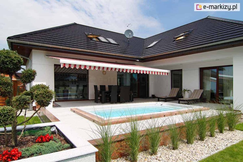 Szeroka, klasyczna markiza tarasowa Palladio Selt w klasyczne, pionowe pasy w kolorze biało-czerwonym, rozłożona nad tarasem luksusowego domu jednorodzinnego. Poszycie wykończone jest dekoracyjną, pofalowaną falbaną z lamówką, która zwisa z przedniego profilu konstrukcji. Markiza jest zamontowana do białej elewacji parterowego budynku o ciemnym, wielospadowym dachu pokrytym dachówką. Pod osłoną znajduje się strefa jadalniana z dużym, ciemnym stołem i kompletem krzeseł. Bezpośrednio przed tarasem zaprojektowano nowoczesną strefę rekreacyjną z wbudowanym w podłoże basenem o jasnych brzegach. Wokół basenu rozciąga się jasna nawierzchnia z płyt tarasowych, przy której ustawiono dwa wygodne leżaki. Ogród jest starannie wypielęgnowany, z ozdobnymi krzewami formowanymi w kule, pasem ozdobnych traw posadzonych w jasnym żwirze oraz czerwonymi kwiatami w nowoczesnej donicy. Całość kompozycji uchwycona w pełnym słońcu podkreśla wypoczynkowy charakter przydomowej przestrzeni.