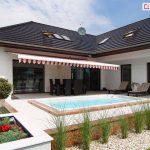 Szeroka, klasyczna markiza tarasowa Palladio Selt w klasyczne, pionowe pasy w kolorze biało-czerwonym, rozłożona nad tarasem luksusowego domu jednorodzinnego. Poszycie wykończone jest dekoracyjną, pofalowaną falbaną z lamówką, która zwisa z przedniego profilu konstrukcji. Markiza jest zamontowana do białej elewacji parterowego budynku o ciemnym, wielospadowym dachu pokrytym dachówką. Pod osłoną znajduje się strefa jadalniana z dużym, ciemnym stołem i kompletem krzeseł. Bezpośrednio przed tarasem zaprojektowano nowoczesną strefę rekreacyjną z wbudowanym w podłoże basenem o jasnych brzegach. Wokół basenu rozciąga się jasna nawierzchnia z płyt tarasowych, przy której ustawiono dwa wygodne leżaki. Ogród jest starannie wypielęgnowany, z ozdobnymi krzewami formowanymi w kule, pasem ozdobnych traw posadzonych w jasnym żwirze oraz czerwonymi kwiatami w nowoczesnej donicy. Całość kompozycji uchwycona w pełnym słońcu podkreśla wypoczynkowy charakter przydomowej przestrzeni.